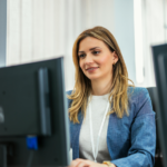 A woman with long hair sits at a desk, focused on her computer screen, dressed in a casual blazer and shirt, with warm, natural lighting.