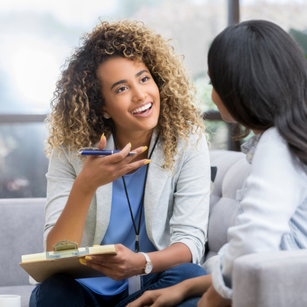 Two women engaged in a conversation on a sofa, one holding a clipboard and pen, surrounded by natural light and modern decor.
