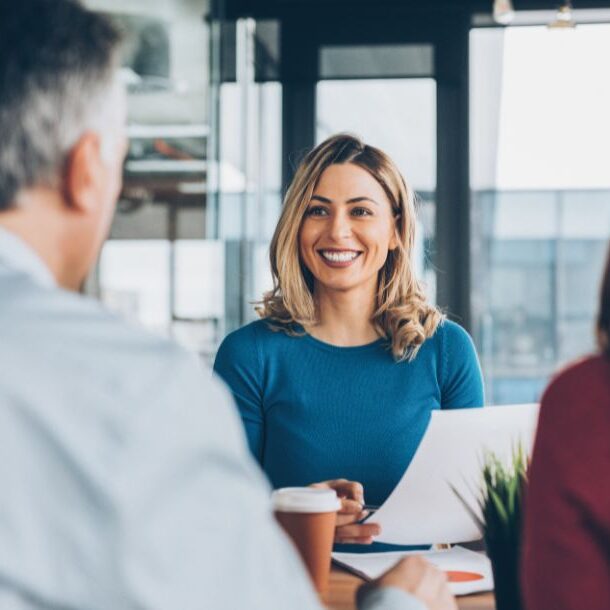 A woman in a blue sweater sits at a conference table, holding documents, with two colleagues listening attentively in a modern office setting.